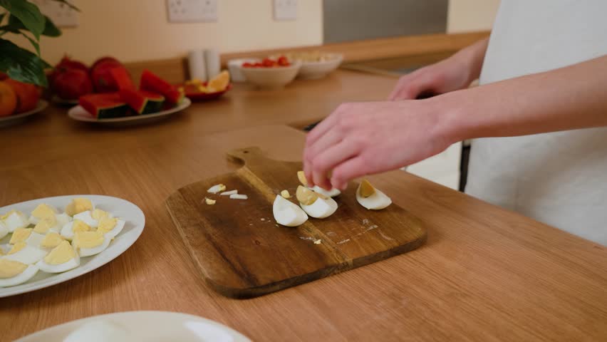Hands placing sliced boiled eggs on wooden cutting board. Person arranging egg pieces during meal preparation. Fingers carefully handling eggs for kitchen task. Hands organizing food for simple
