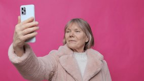 Smiling older woman enjoys a moment capturing a selfie. She stands in front of a vivid pink backdrop, wearing a cozy sweater and soft outerwear. Her joyful expression reflects confidence - Powered by Shutterstock - Get 15% off with code: PIKWIZARD15