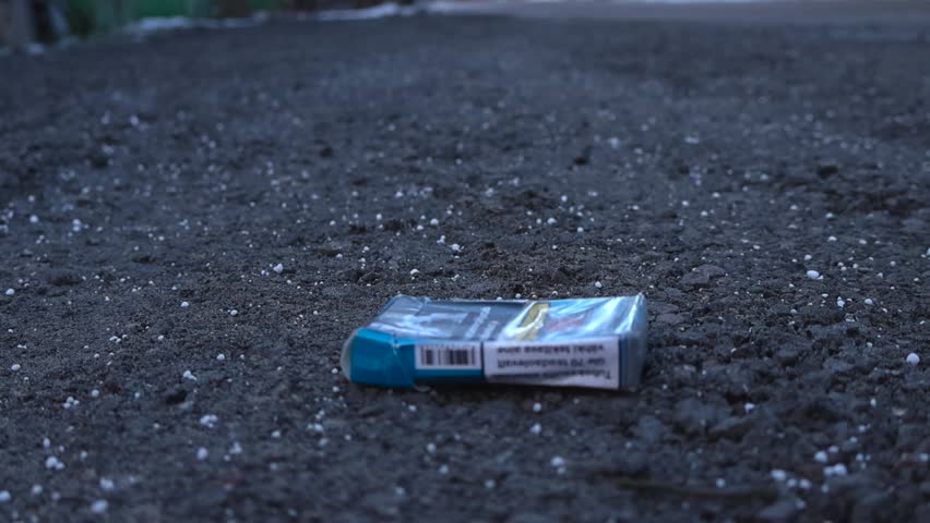 Close up left to right panning footage of a blue and wrinkled cigarette pack that is on a dark gravel and muddy ground during a cloudy day. Some snow grains are visible in the ground. In focus smokes