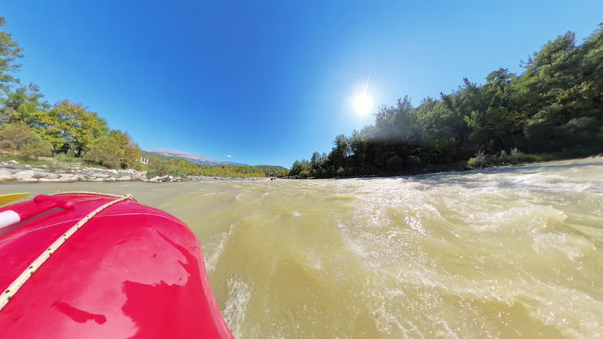 A head-on view of a splash hitting a rafting boat as it tackles fast-moving rapids under a clear blue sky, with paddles visible and water droplets flying through the air.