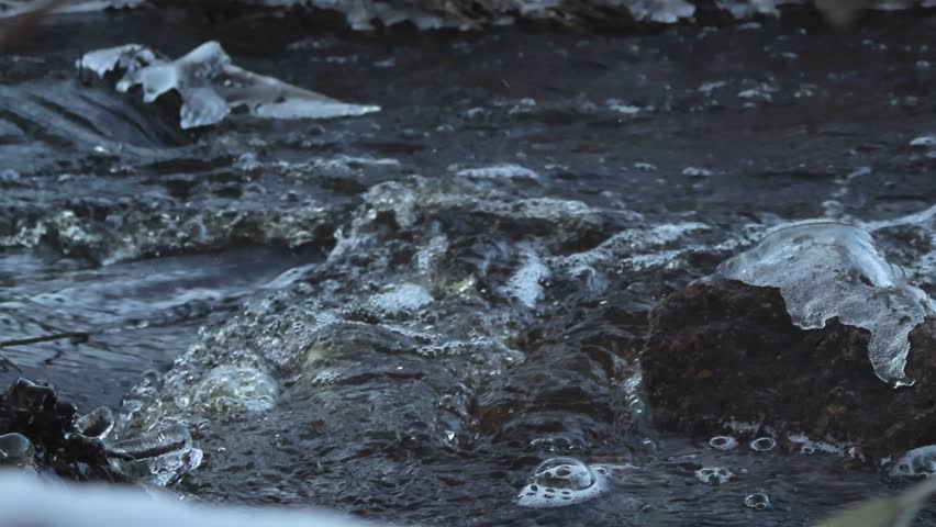 Close up slow motion footage of dark river water flowing during winter day with foamy and bubbly waves. Ice covered rocks are in the water and water is in sharp focus. Snowy river bank visible in back