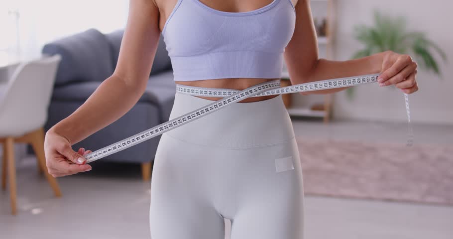 Cropped portrait of faceless slim sporty woman wearing sportswear measure the waist with measuring tape standing at home. Slimming, sport, diet and weight control concept. 4k video.
