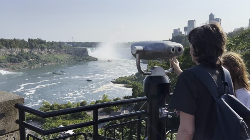 Visitors using telescopes to view the majestic Niagara Falls on a sunny day, surrounded by lush greenery and city skyline, Canada