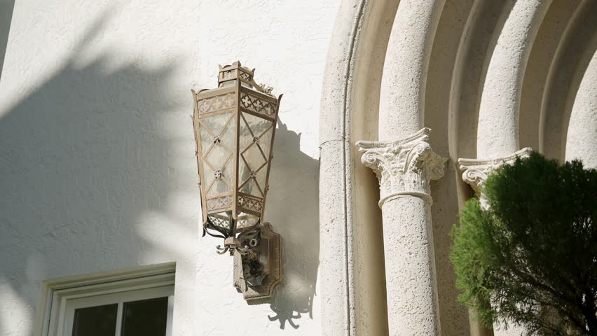 Exterior wall of building with decorative lantern attached to it. Lantern has vintage design with intricate metalwork and glass panels. Wall is textured and painted white