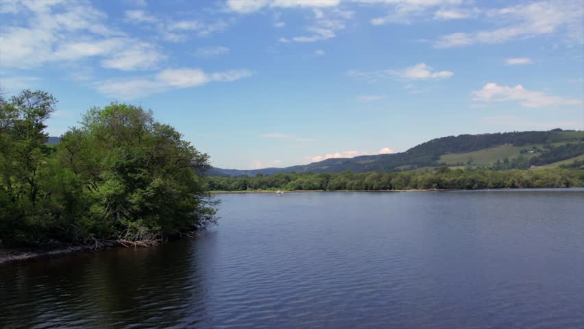Loch Ness shoreline with lush green forests and calm water reflecting the vibrant blue sky. Captures the picturesque scenery and iconic landscape of the Scottish Highlands.