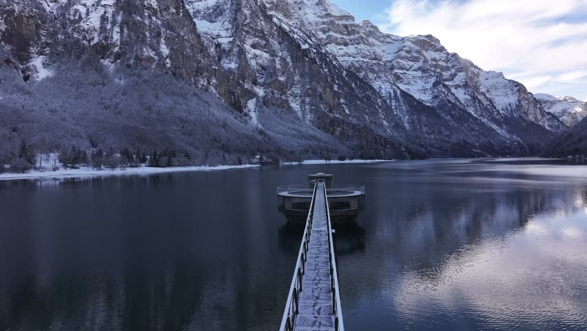 Winter scene at Klöntalersee, Switzerland. Snow-capped mountains frame the crystal-clear lake. A unique circular spillway structure adds a touch of modern engineering to the natural beauty.