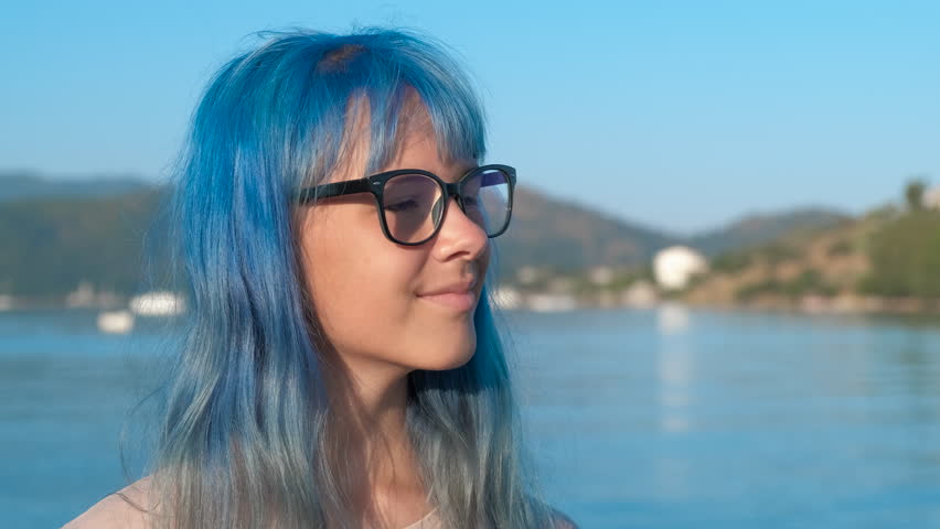 Beautiful stylish teen against river. Cute girl with blue hair and eyeglasses on the background of the blue sea.