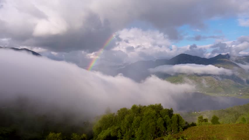A breathtaking view of a misty mountain landscape, with clouds rolling over lush green valleys and a shimmering lake below. A vibrant rainbow arches across the sky, adding a magical touch.