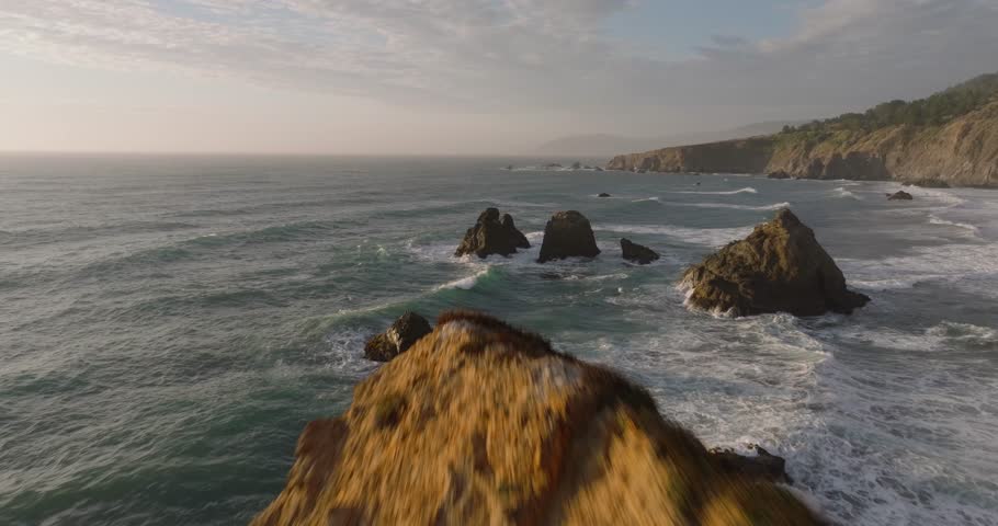 Drone fast pullback over sea stack ridge along Highway 1 in California with ocean waves and dramatic cliffs under a clear twilight golden sky