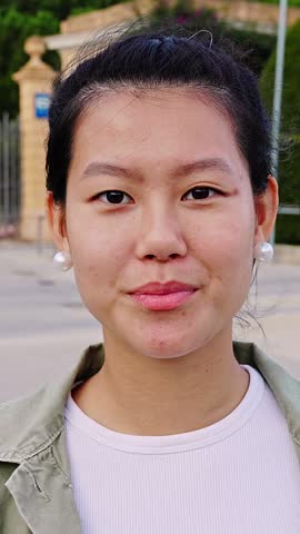 Vertical HD video. Happy young asian teen girl smiling at camera standing at city street. Outside portrait of joyful Chinese woman over urban background.