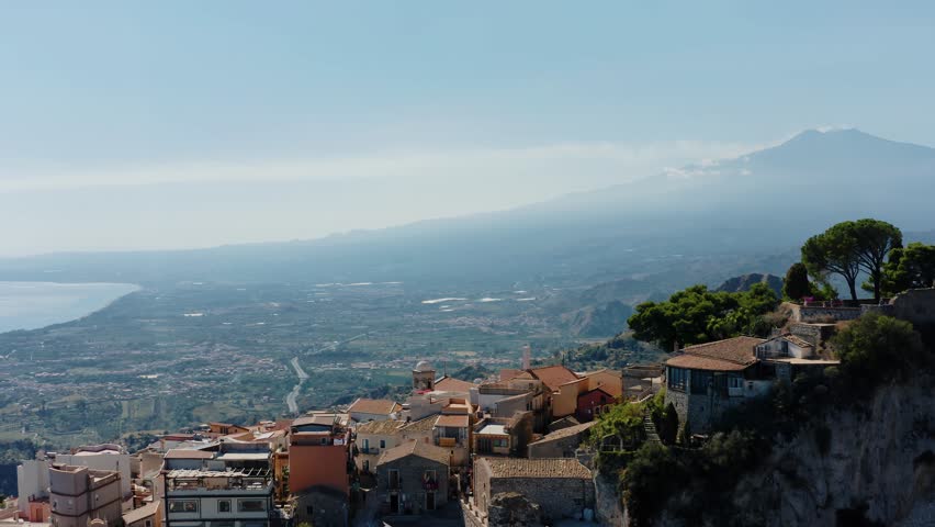Drone shot flying towards Mount Etna with buildings of Castelmola town below. Historic town high up in the mountains in Sicily. Panoramic view to Mediterranean sea and volcano.