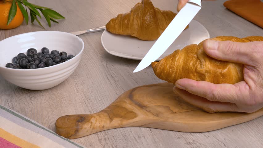 A man cuts croissants with a knife on a wooden cutting board.