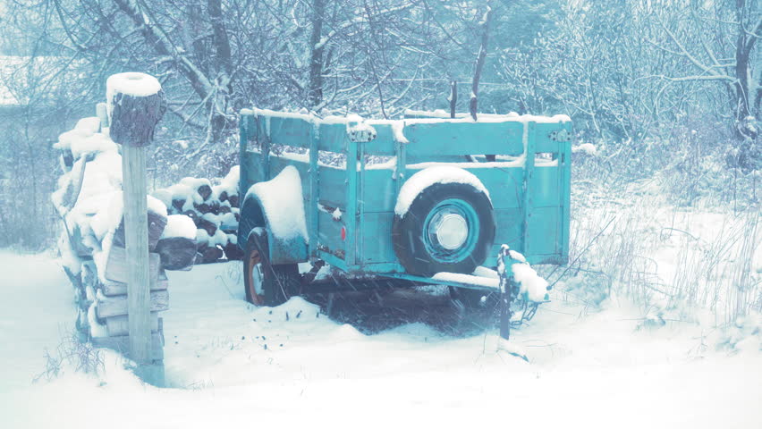 Old wood trailer with chipped blue paint surrounded by snow covered chopped wood and tree in distance during a heavy snow