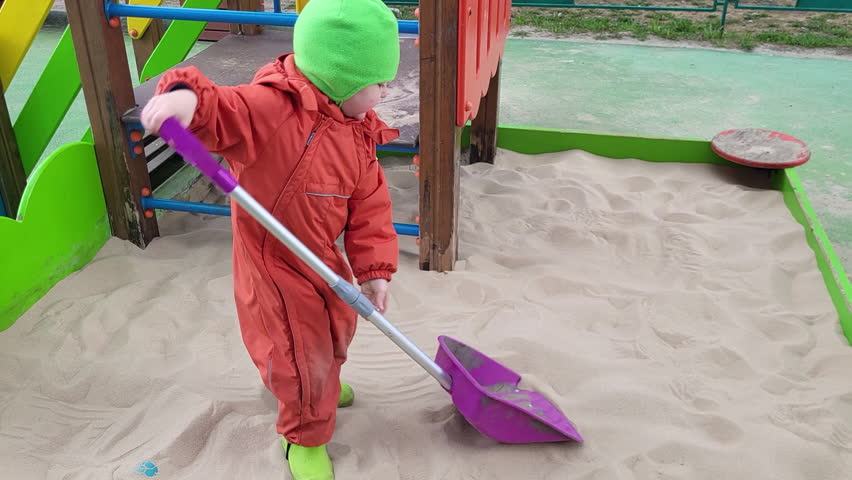 Child in orange snowsuit playing in sandbox with purple shovel. Outdoor playground scene. Winter clothing and activity concept.