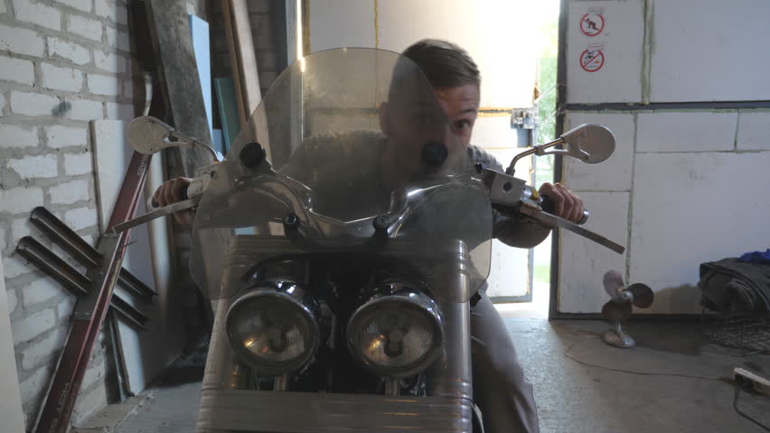Young man pretending to fast ride a moto in workshop. Cheerful guy fooling around sitting on motorcycle in shed. Happy human enjoying and having fun in garage. Close up Slow motion