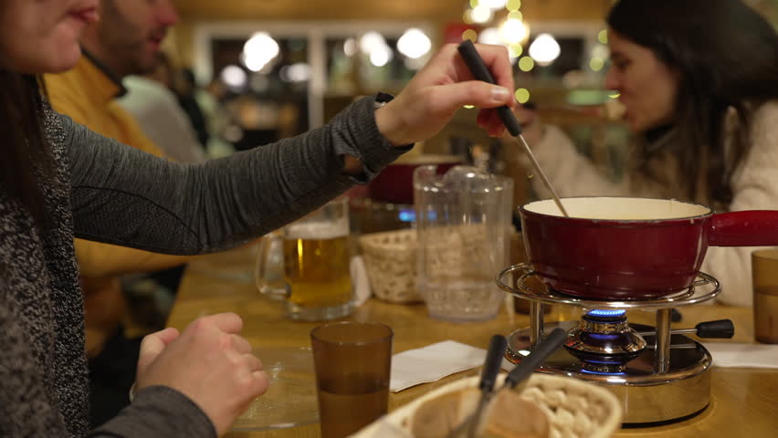 Closeup of Cheese Fondue Dining - Hand Dipping Bread in Melted Cheese, Red Pot Over Heater in Restaurant