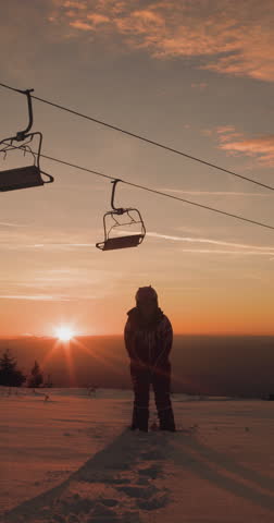 Silhouette of a female skier against sunset sky beneath chairlift. Girl jumping and throwing snow. 120fps cinema camera