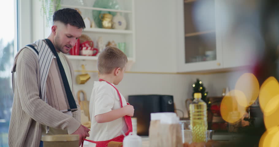 A dad ties an apron for his young son, and they begin baking Christmas gingerbread cookies together in the kitchen.