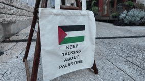 Canvas tote bag displayed on a wooden stand in an urban setting, featuring the Palestinian flag and the text - Keep Talking About Palestine. Concept of activism, solidarity, and freedom of expression - Powered by Shutterstock - Get 15% off with code: PIKWIZARD15