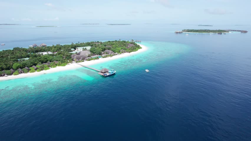 A drone view of an island with white sand in the middle of the ocean. Turquoise water, coral reef. There are bungalows on the water and villas on the beach. There are palm trees all around. Maldives