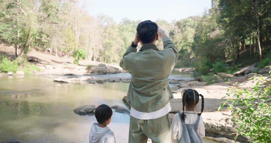 Asian single father standing with two children by the river in a forest, using binoculars to explore wildlife, fostering family bonding, love, and appreciation for nature’s beauty.