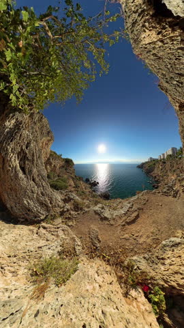Vertical video. Wide Angle. A panoramic view from inside a natural rock formation overlooking the sunlit sea. The rugged cliffs and greenery frame the calm ocean, creating a beautiful and peaceful coa