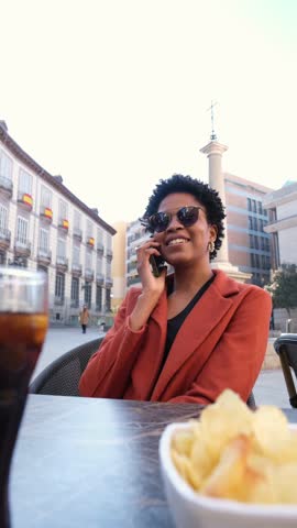 Happy businesswoman making a phone call while enjoying chips and a drink at an outdoor cafe in a square in Madrid