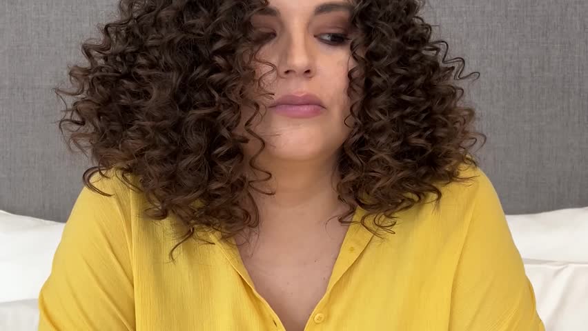 Horizontal video close up. Young adult white sad child free brunette woman with curly hair wearing yellow shirt, party hat and blowing vuvuzela in hotel room. Loneliness, holiday, depression, joy, fun