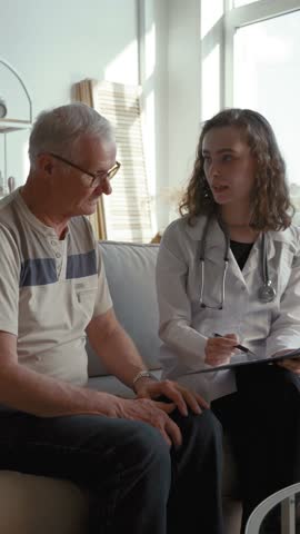 Lady doctor in white coat prescribes medications for elderly patient at home. Specialist shows treatment plan to senior man sitting on sofa in living room Vertical Shot.