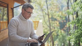 Mature businessman using laptop and smartphone on wooden terrace of mountain chalet, remote working from peaceful nature. Portrait of happy mid adult man wit grey hair and glasses, smiling. - Powered by Shutterstock - Get 15% off with code: PIKWIZARD15
