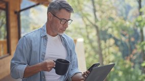 Mature man drinking morning coffee and using laptop on terrace of wooden cabin in mountains, enjoying remote work and freedom. Entrepreneur businessman managing business online from nature. - Powered by Shutterstock - Get 15% off with code: PIKWIZARD15