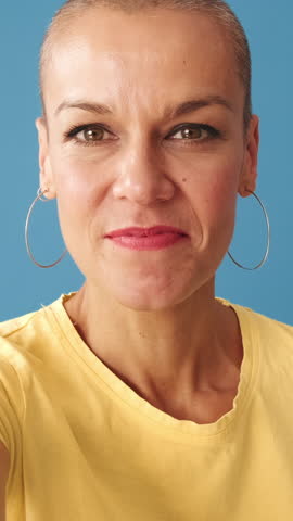 Vertical video, Close-up of young bald woman wearing yellow t-shirt looking at the camera video call, waving hand in greeting gesture, isolated on blue background in studio