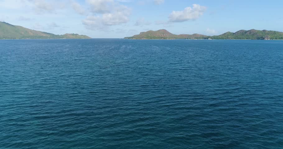 Aerial rocky island sits in the middle of the ocean. The water is calm and clear, and the sky is blue. The scene is peaceful and serene