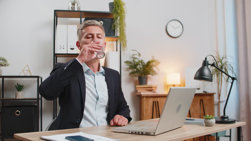 Focused Caucasian businessman sitting at workplace desk drinking water while working with laptop in home office. Stylish young employee freelancer in suit looks at computer screen browse surf internet