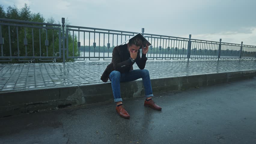A man is sitting on the edge of a sidewalk, sheltering his head from the rain while looking pensive. The grey sky adds to the atmosphere of solitude in the city.