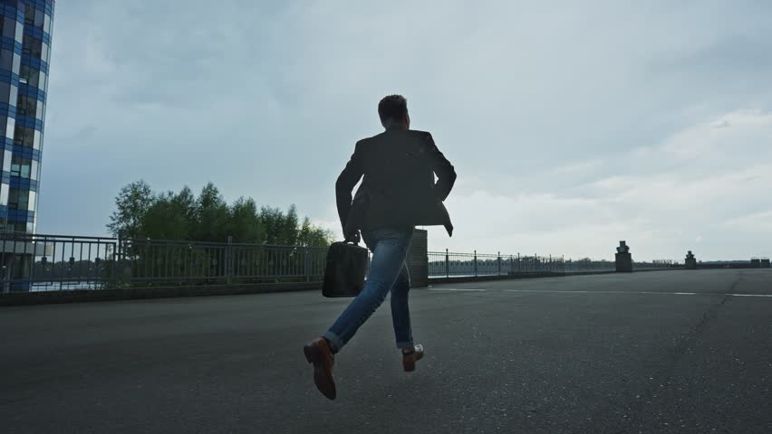 A person hurries along a waterfront walkway under overcast skies in the late afternoon. The atmosphere suggests an impending rain, creating a sense of urgency and motion.