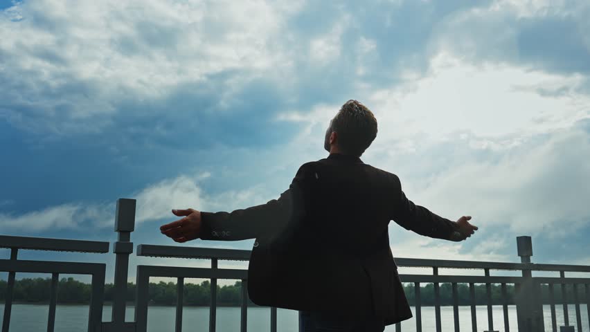 A man stands by the water, arms wide open, embracing the tranquility of the moment as clouds drift overhead. The serene atmosphere invites a sense of calm and reflection.
