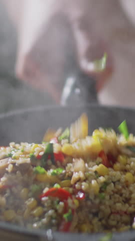 Vertical close-up shot of hands of unrecognizable chef stir-frying steaming rice and vegetables in hot pan while cooking food