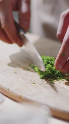 Vertical close-up shot of hands finely chopping fresh green herbs on wooden cutting board using sharp knife highlighting preparation of aromatic ingredients
