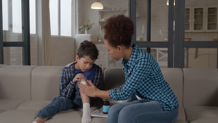 Loving pretty black mother treating injured knee of African American preadolescent son after fall , applying adhesive band aid on child's leg in domestic room.