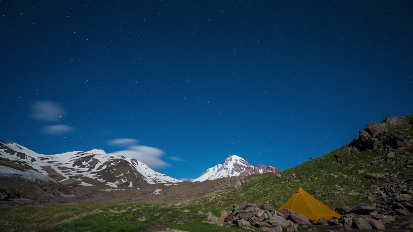 Night timelapse of a tent under starry sky with Kazbegi peak in background, Caucasus mountains. Stars move across sky. Remote camping, nature adventure, eco tourism. Peaceful wilderness.
