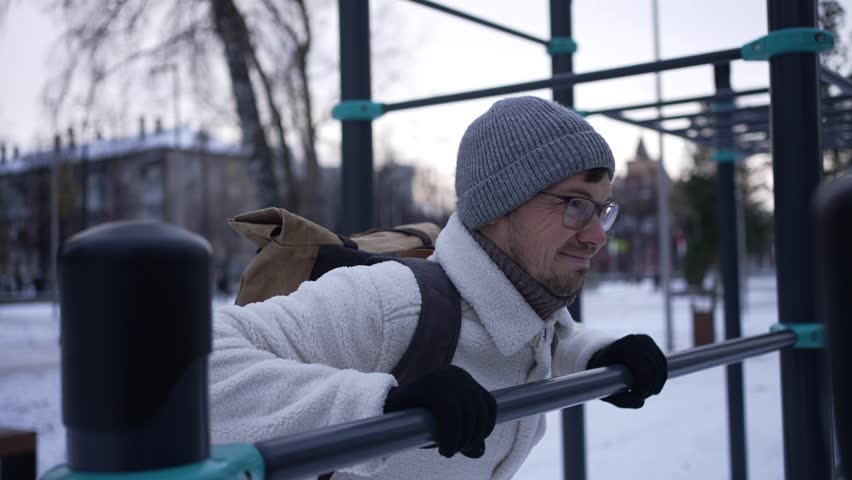 Determined man wearing a warm winter jacket, beanie, and gloves performs dips on parallel bars in a snow-covered park, demonstrating dedication to fitness despite the cold weather