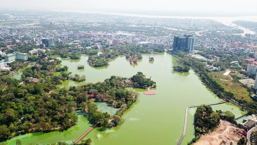 A stunning aerial perspective of Kandawgyi Lake in Rangun, Myanmar, showcasing its lush green surroundings, small islands, and the vibrant cityscape in the background