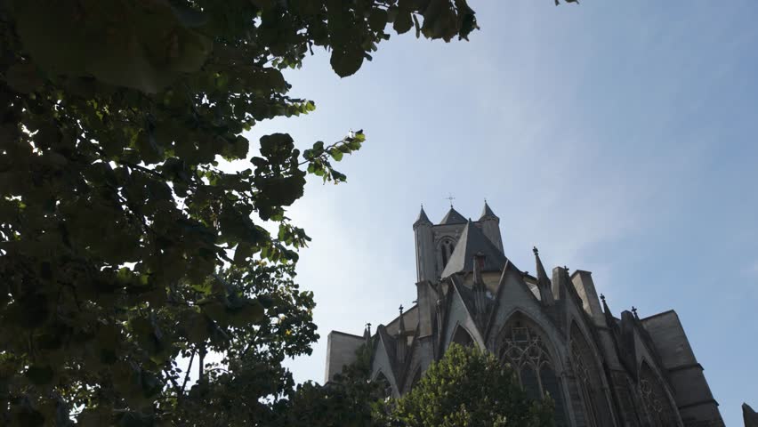 View of St. Nicholas Church in Ghent, Belgium, surrounded by greenery under a clear blue sky. The Gothic architecture harmonizes with the serene trees
