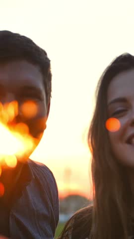 The couple dancing with firework sticks on an morning sunrise background. Super slow motion. Close-up of fireworks and sparklers. Re-focusing from sparklers to faces. Vertical shot.