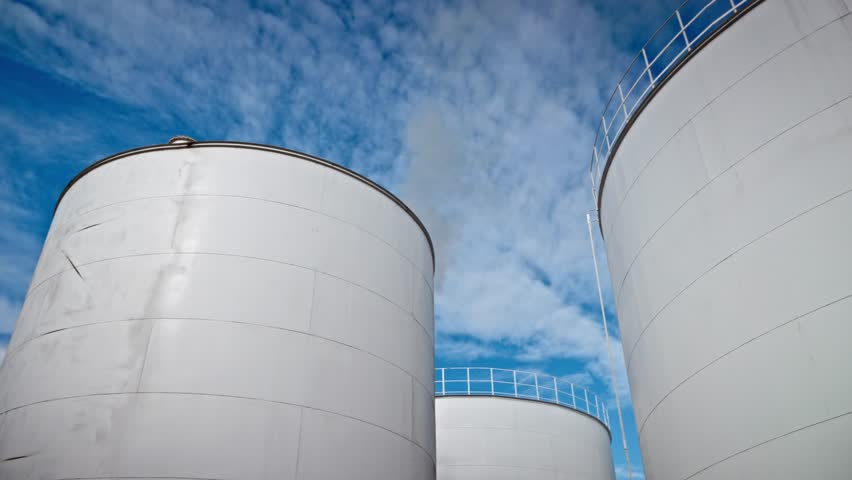 Large industrial storage tanks under a bright blue sky with scattered clouds. The white cylindrical structures feature metal railings on top, standing out against the vibrant backdrop.