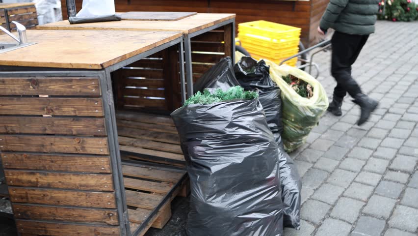 Large black and green plastic bags filled with dismantled Christmas decorations are placed near a wooden counter as the cleanup process continues. A person walks by on a cobblestone street, signaling 