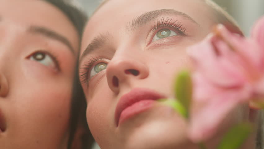 Two beautiful skin care faces multiracial girls women female Caucasian Asian Korean Chinese Japanese indoors holding flowers delicate facial surgery aromatherapy natural eco cosmetic gentle perfume