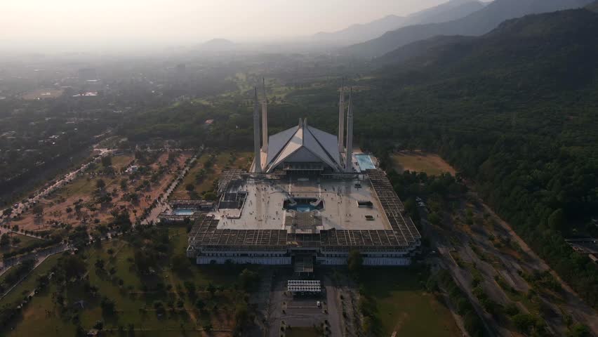 Aerial view of the majestic Shah Faisal Mosque surrounded by serene mountains and lush greenery at sunset, Islamabad, Pakistan.