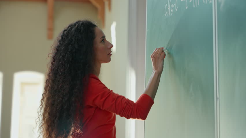 Smiling clever Hispanic student girl Latina woman Caucasian female lady teacher tutor studying learn writing chalk on blackboard formula math mathematic in class auditorium university college academy
